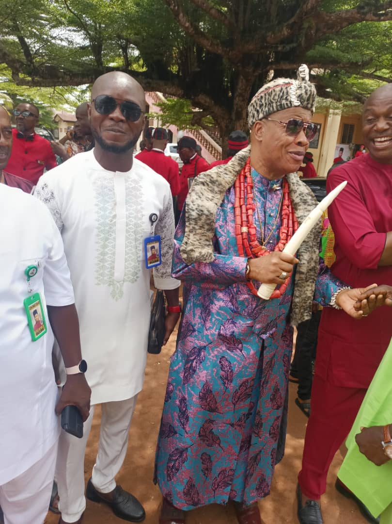 A man in a feathered cap and red beads holding a carved ivory-coloured horn, with people crowded around him