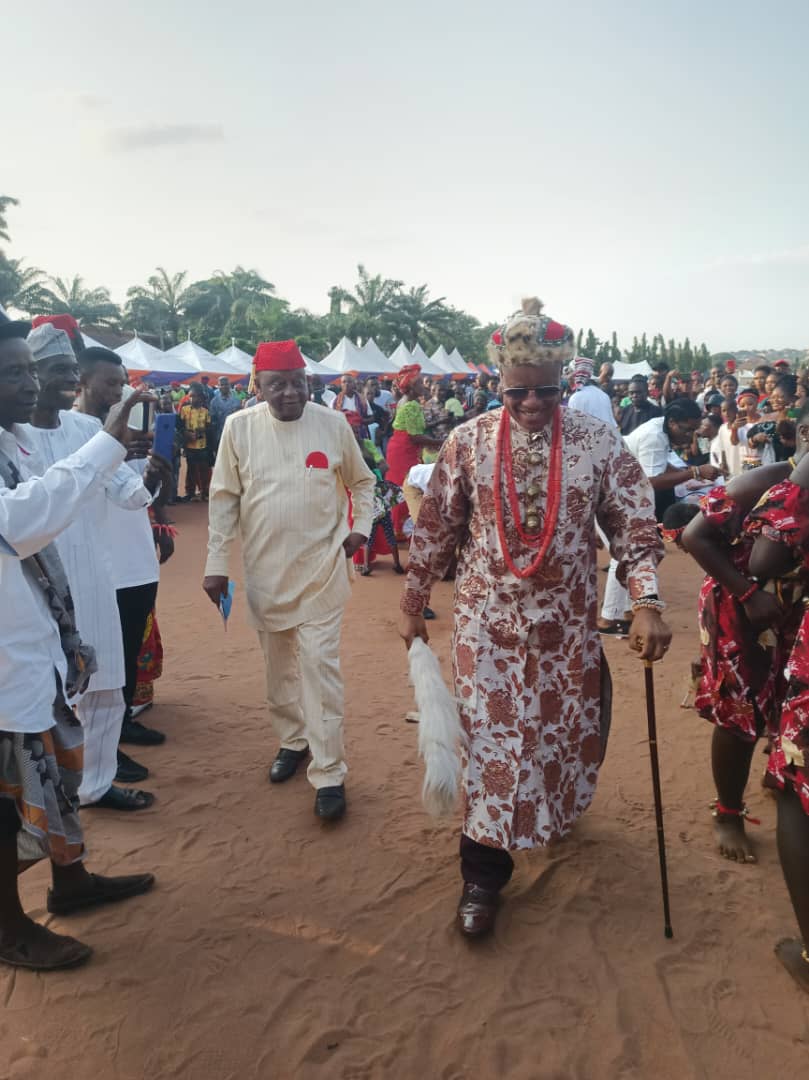 A community leader in red-patterned regalia walking with a cane through a crowd
