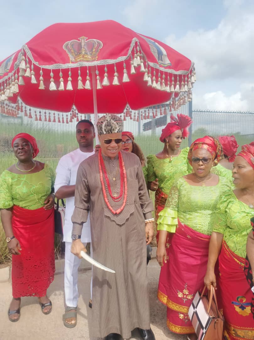 A leader in traditional dress holding a ceremonial sword beneath a large red umbrella, surrounded by women in matching green and red outfits