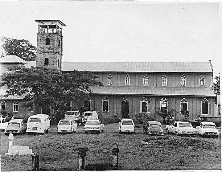 Archival black-and-white photograph of a long church with a bell tower and vintage cars parked in front